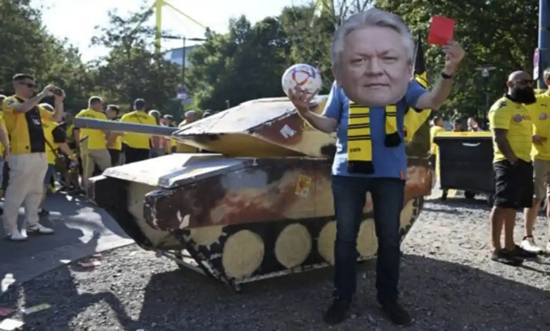 Un tanque ficticio junto a un activista que lleva una máscara con la imagen del director ejecutivo de Rheinmetall, Armin Papperger, y sostiene una tarjeta roja antes de un partido de fútbol en Dortmund, Alemania, el 24 de agosto de 2024. Con el impulso hacia la guerra y el mayor gasto en armas, los directores ejecutivos de armamento como Papperger están esperando que sus ganancias exploten. | Bernd Thissen / dpa vía AP